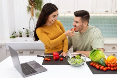 Güzel, esmer, uzun saçlı, yakışıklı erkek arkadaşını besleyen, birlikte yemek yapan, dizüstü bilgisayarda yemek videosu izleyen, aşıklar yemek pişirirken eğlenen bir kadın.