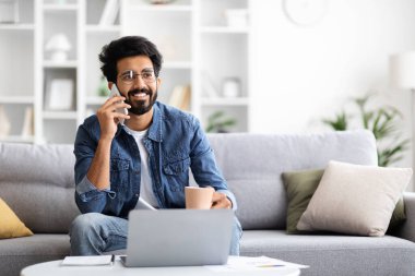 Mutlu Hintli serbest çalışan adam cep telefonuyla konuşuyor, dizüstü bilgisayar kullanıyor ve koltukta otururken kahve içiyor, gülümseyen doğulu adam mobil iletişimin keyfini çıkarıyor,