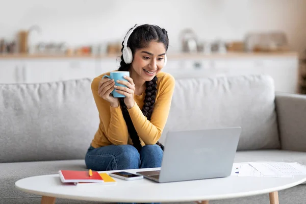 Cheerful indian woman watching videos on laptop and drinking coffee at home, beautiful happy eastern female wearing wireless headphones looking at computer screen and smiling, copy space