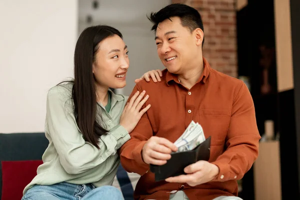 給与と貯金の概念 幸せな韓国の結婚した配偶者は お札を財布に入れて お互いを見て笑って 自宅でソファに座っています — ストック写真
