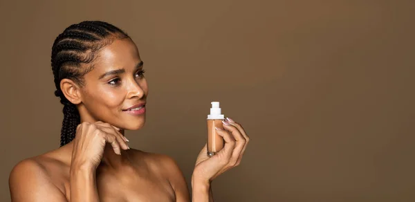 黑色中年妇女拿着装有液体粉底的瓶子 看着空余的空间为您的广告 每日化妆的女性 站在褐色背景上 — 图库照片