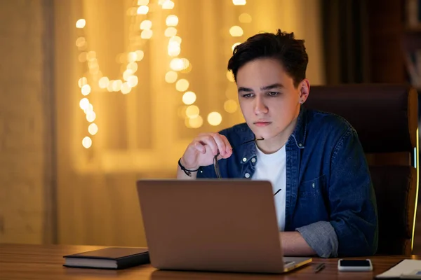 Pensive handsome young caucasian guy in casual outfit sitting at workdesk, holding eyeglasses, looking at computer screen, developer working on laptop at office late at night, copy space