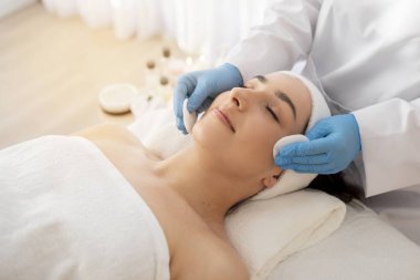 Kozmetik uzmanı, pamuk pedli genç Hintli kadının derisini temizliyor, modern spa salonunda rahatça yüz bakımı yapıyor, güzellik salonunda sağlık günü düzenliyor.