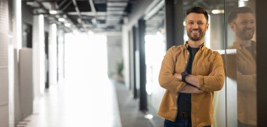 Ofiste ya da fabrikanın içinde kollarını çapraz bağlayan mutlu, kendinden emin Avrupalı genç iş adamı. Güneş ışığı, fotokopi alanı. Profesyonel ve iş dünyası, patron, liderlik ve CEO.