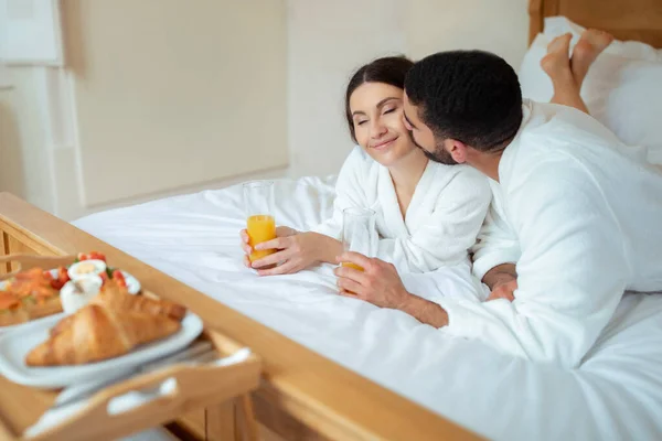 浪漫的早餐 丈夫亲吻妻子谢克喝着果汁躺在靠近盘子的地方 在现代酒店房间的房间里吃着放松的饭 不同的配偶一起享受假期 — 图库照片