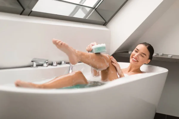 泡でお風呂に入る魅力的な女性 バスルーム内の白い浴槽で肌にシャワージェルを適用スポンジで足を洗う 体をケアする女性の入浴 選択的フォーカス — ストック写真