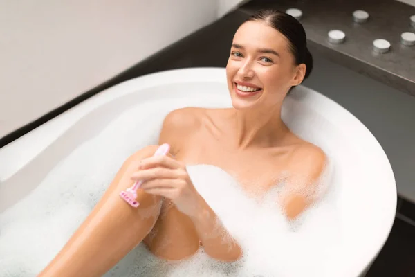 以上是快乐女人用安全剃须刀刮腿的景象 在家里的现代化浴室里 她用泡沫坐在水里拍照 体格护理和头发摘除 — 图库照片