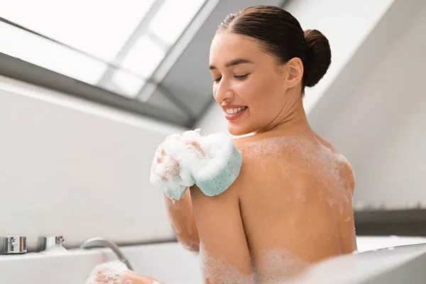現代の浴室の内湯に座っているスポンジゴム製の肩を使って泡と洗濯機でお風呂に入る女性 美容と衛生化粧品のコンセプト 選択的フォーカス — ストック写真
