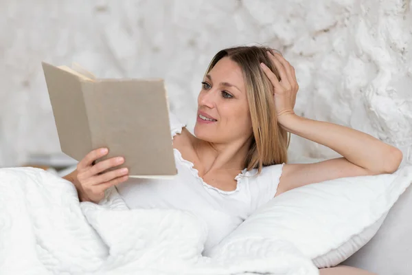 国内服で笑顔スマート千年紀白人ブロンドの女性はベッドの上にあり 良い朝 平和を楽しんで 白い寝室のインテリアで本を読み取ります 自由時間に趣味 休息とリラックス — ストック写真