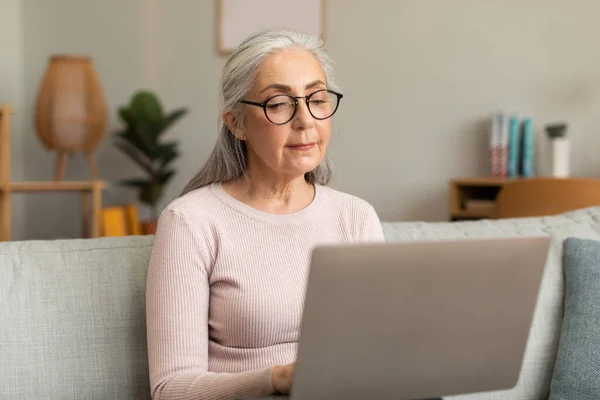Concentrated serious caucasian old lady in glasses with laptop works online, watch video lesson, blog, surfing in internet in living room interior. Study, business, social distance with device at home