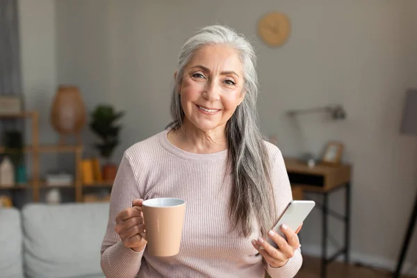 快乐的高加索老年女性 带着智能手机和酒杯 享受着闲暇时间 在客厅里聊天 咖啡休息时间 社交媒体应用 在家里上网的装置 — 图库照片