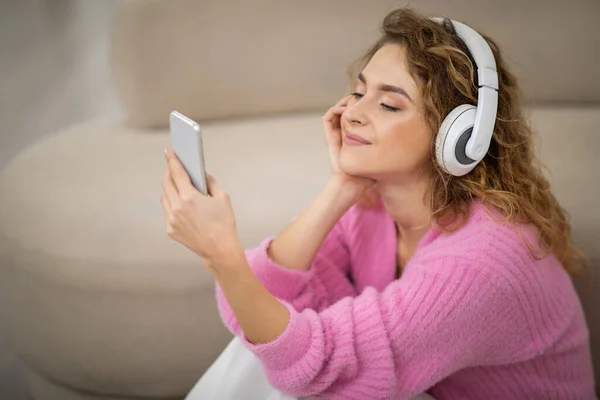 好きなレジャー 美しい若い女性は自宅でスマートフォンを使用してワイヤレスヘッドフォンを身に着けています 魅力的な千年紀の女性は 音楽を聴いたり 携帯電話でインターネットを閲覧 — ストック写真