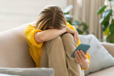 Mutsuz, umutsuzca ağlayan bin yıllık beyaz, akıllı telefonlu, depresyon, yalnızlık ve oturma odasındaki kanepede ayrılık acısı çeken sarışın kadın. Kötü haberlere tepki, evde duygular