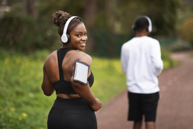 Gülümseyen milenyum Afro-Amerikan çift spor giyim, kablosuz kulaklıklar açık havada, arkada akıllı telefonlarla çalışıyor. Müzikle kilo kaybı, birlikte koşu, spor eğitimi, vücut bakımı.