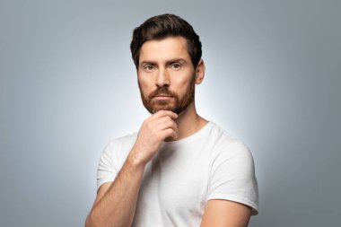 Çekici, bakımlı orta yaşlı bir adamın portresi. Çeneye dokunuyor ve kameraya bakıyor. Gri renkli arka planda izole bir şekilde poz veriyor.