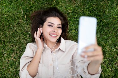 Bin yıllık beyaz kulaklıklı bayan öğrencinin çimenlerin üzerinde yatıp üniversitede ve dışarıda telefonla selfie çekmesine sevindim. Çalışmayı bırak, blog ve sosyal medya için fotoğraf çek, yaşam tarzını değiştir ve rahatla