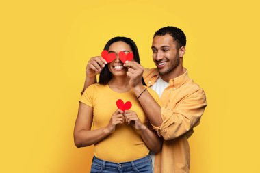 Bin yıllık Afro-Amerikan erkeğinin eşinin gözlerine kalp koymasına, sarı stüdyo arka planında izole edilmiş eğlenmesine sevindim. Tatil, yıldönümü, Sevgililer Günü, aşk ve romantizm için tebrikler.