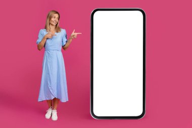 Orta yaşlı Avrupalı sarışın kadın boş ekranlı büyük bir telefonu işaret ediyor. Tavsiye uygulaması, pembe stüdyo arka planında izole edilmiş bir web sitesi. Sunum, aygıt önerisi, sohbet