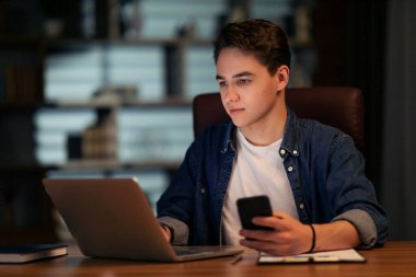 Genç yakışıklı adam ofiste geç saatlere kadar çalışıyor, çalışma masasında oturuyor, modern laptop ve akıllı telefon kullanıyor, aylık rapor üzerinde çalışıyor, iş telefonu uygulaması kullanıyor, yer kopyalıyor.