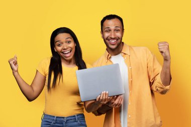 Laptopları ve ağzı açık, zafer ve başarı jestleri yaparak sarı arka planda izole edilmiş binlerce yıllık Afro-Amerikan çifti heyecanlandırdığına sevindim. Online alışveriş, satış ve kazanç