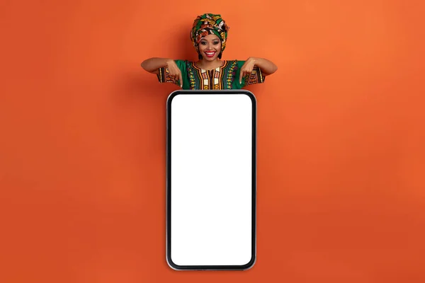 興奮した幸せな陽気なかなりアフリカの若い女性指している巨大なスマートフォンで空の白い画面 黒の女性を身に着けている民族衣装 オレンジのスタジオの背景 モックアップ コピースペース — ストック写真