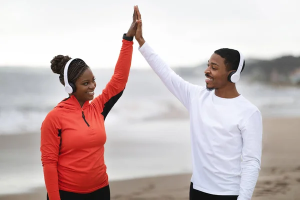 Cheerful millennial african american couple in sportswear and headphones giving high five, celebrating great run on beach outdoor. Break, rest from jogging together, sports, training and body care