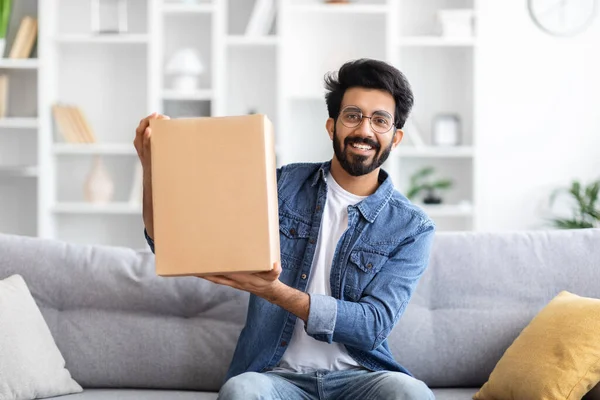 Handsome Young Indian Man Demonstrating Cardboard Box With Delivery While Sitting On Couch At Home, Eastern Guy Holding Delivered Parcel In Hands And Smiling At Camera, Enjoying Fast Shipping