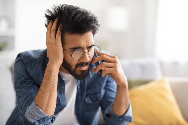 Worried Young Indian Man Talking On Cellphone At Home, Stressed Millennial Eastern Guy Having Unpleasant Mobile Conversation, Got Bad News, Touching Head And Looking Away, Closeup Portrait