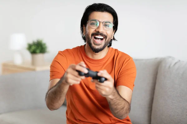 ひげを生やした眼鏡で幸せな千年紀のアラビア人の男性ゲーマーはジョイスティックでソファに座って リビングルームのインテリアで一人でオンラインゲームをプレイします ライフスタイル 自宅での楽しさとエンターテイメントのためのガジェット — ストック写真