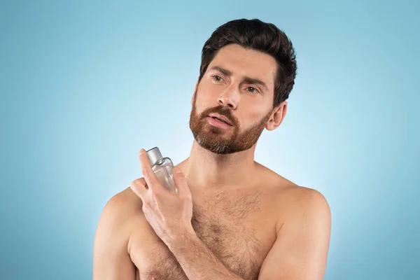 Bearded middle aged man spraying on cologne, doing morning routine over blue studio background. Beauty skin care, moisturizers products and perfume