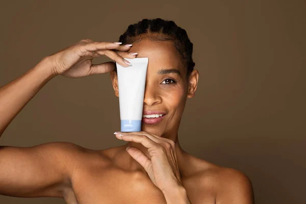 アフリカ系アメリカ人の中年女性が化粧品クリームでチューブを保持笑顔 目をカバー 茶色のスタジオの背景にポーズ コピースペース 美容製品コンセプト — ストック写真