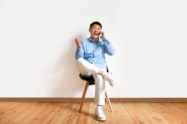 Возбужденный Корейский Мужчина Разговаривает Смартфону Сидит Стуле Фоне Белой Стены — стоковое фото