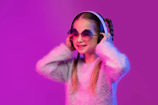 甘いです可愛いです可愛いですブロンド女の子学校 サングラスで現代のワイヤレスヘッドフォンを使用して 幸せな子供は音楽を聞いて 楽しみますピンクのスタジオの背景にネオンライト — ストック写真