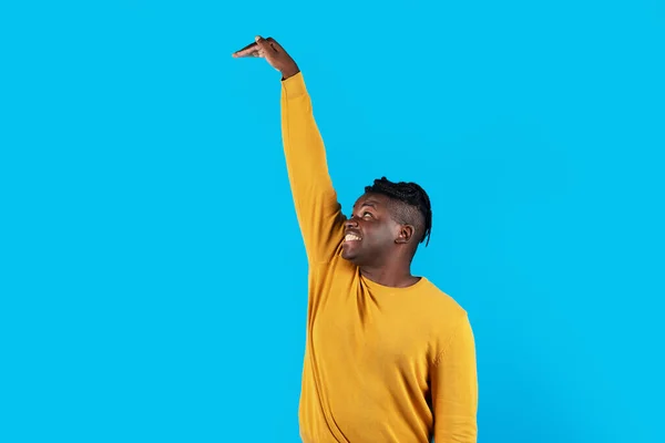Cheerful African American Guy Measuring High Invisible Object With Hand While Standing Isolated Over Blue Studio Background, Young Black Man Looking Up And Checking Hight, Copy Space