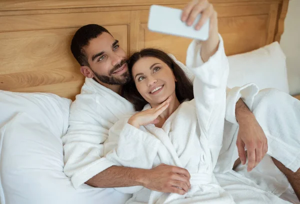 ホテル内のベッドルームにある白いバスローブを着た自撮りポーズをとるスマートフォンを使った幸せなカップル 休暇を楽しんで写真を撮る新婚旅行を楽しんでいる配偶者 選択的フォーカス — ストック写真
