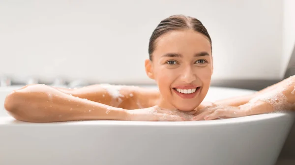 自宅の近代的なバスルームでカメラに笑みを浮かべて浴槽にポーズを取る豪華な女性 ソープスッドで覆われた体のための女性の入浴ケア 美容ルーチンのコンセプト パノラマ — ストック写真