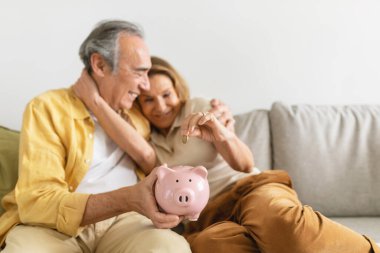 Aile birikimi. Mutlu kıdemli çift evdeki kanepede birlikte otururken kumbaraya para koyuyor. Eşlerini seviyorlar, bütçe planlıyorlar ve gelecek için para biriktiriyorlar.
