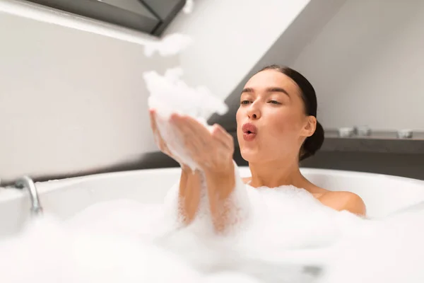 お風呂でお風呂に入る楽しみを持つ泡を吹くリラックスした女性自宅でバスルームに石鹸泡でお湯でいっぱい 衛生ルーチンの入浴室内を楽しむ女性 選択的フォーカス — ストック写真