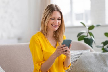 Bin yıllık neşeli, sarı elbiseli beyaz kadın akıllı telefondan mesaj okuyor, sosyal ağlarda sohbet ediyor, boş zamanlarında internette oda içinde sörf yapıyor. Haberler, uygulama, reklam ve teklif