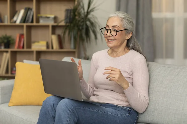 stock image Smiling pretty caucasian aged woman teacher in glasses has online call, gesturing, watch video lesson, shooting vlog in living room interior. Application for communication, meeting, study and tutoring