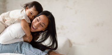 Sevimli Asyalı kız bebek annesine sarılıyor ve evde eğleniyor. Mutlu anne ve yeni yürümeye başlayan bebek yatak odasında gülüp kaynaşıyor. Metin için Boş Alanı Olan Panorama