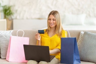 Bin yıllık neşeli beyaz kadın mağazadan bir sürü paketi olan alışverişçi sarışın kadın kredi kartı, dizüstü bilgisayar kullanıyor ve oturma odasında online alışveriş yapmaktan hoşlanıyor. Satış, nakit para, eve sipariş.