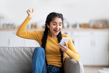 Harika haberlerim var. Coşkulu genç Hintli kadın akıllı telefon ekranına bakıyor ve evdeki koltukta otururken başarıyı kutluyor, Mutlu Doğu kadını heyecanla bağırıyor ve yumruğunu kaldırıyor,