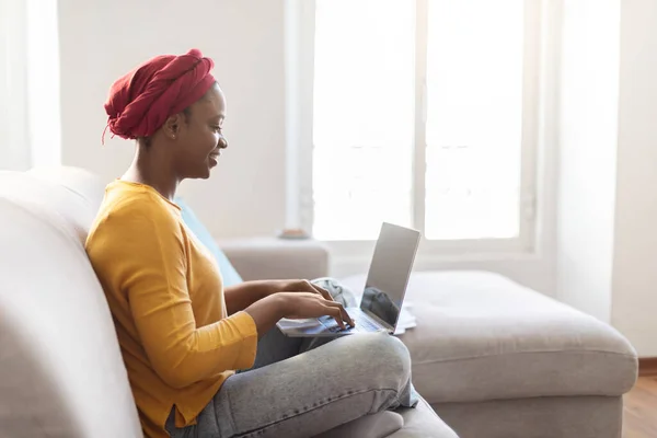 Freelance, remote job. Side view of young stylish black woman wearing casual outfit and traditional turban using laptop with empty blank screen, typing on computer keyboard, mockup, copy space for ad