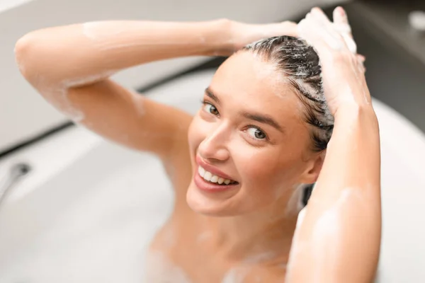 自宅の浴室にある浴槽で頭を洗うカメラにシャンプーの笑顔を適用する魅力的な女性 化粧品でヘアケアを楽しむ幸せな女性の入浴 選択的フォーカス — ストック写真
