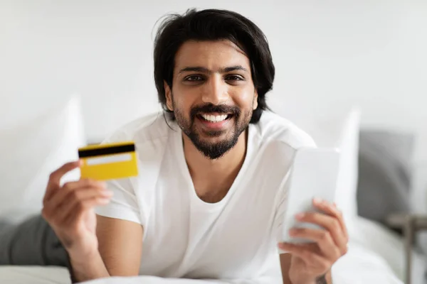 Usmívající Tisíciletý Arabský Muž Plnovousem Leží Posteli Ukazuje Kreditní Kartu — Stock fotografie