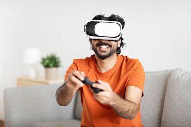 VR gözlüklü, sakallı, kanepeye oturup oturma odasında online oyun oynayan mutlu bin yıllık Arap erkek oyuncu. Yaşam tarzı, evde eğlence ve eğlence aracı