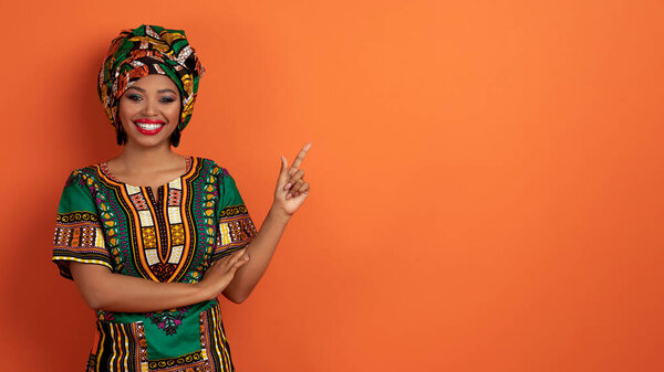 Cheery happy attractive young black woman in green colorful african costume showing nice offer, pointing at copy space for text or advertisement and smiling, isolated on orange background, banner