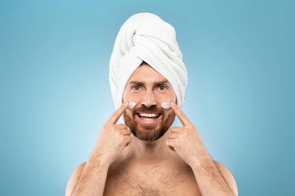 顔に保湿クリームを塗る白人髭男 カメラと笑顔を見て 青いスタジオの背景に頭の上にタオルで立って — ストック写真