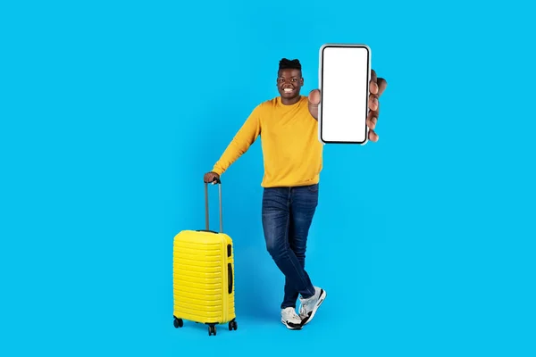 Smiling Handsome Black Man With Suitcase And Blank Smartphone In Hand Standing Over Blue Studio Background, Smiling Young African American Guy Recommending New Travel App, Collage, Mockup